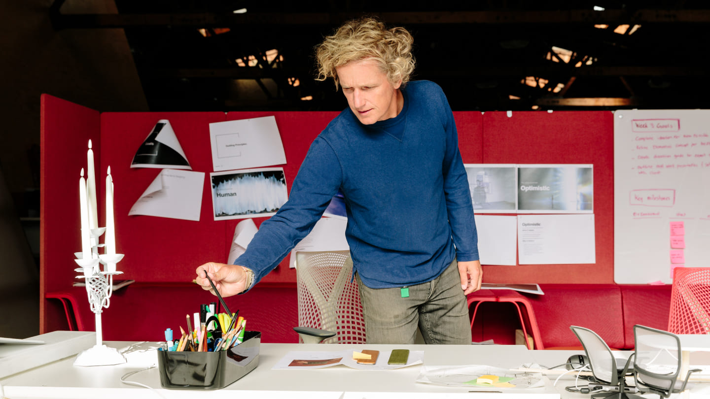 Yves Béhar reaching for a drawing tool at a studio worktable, with prototypes and pinned ideas behind him