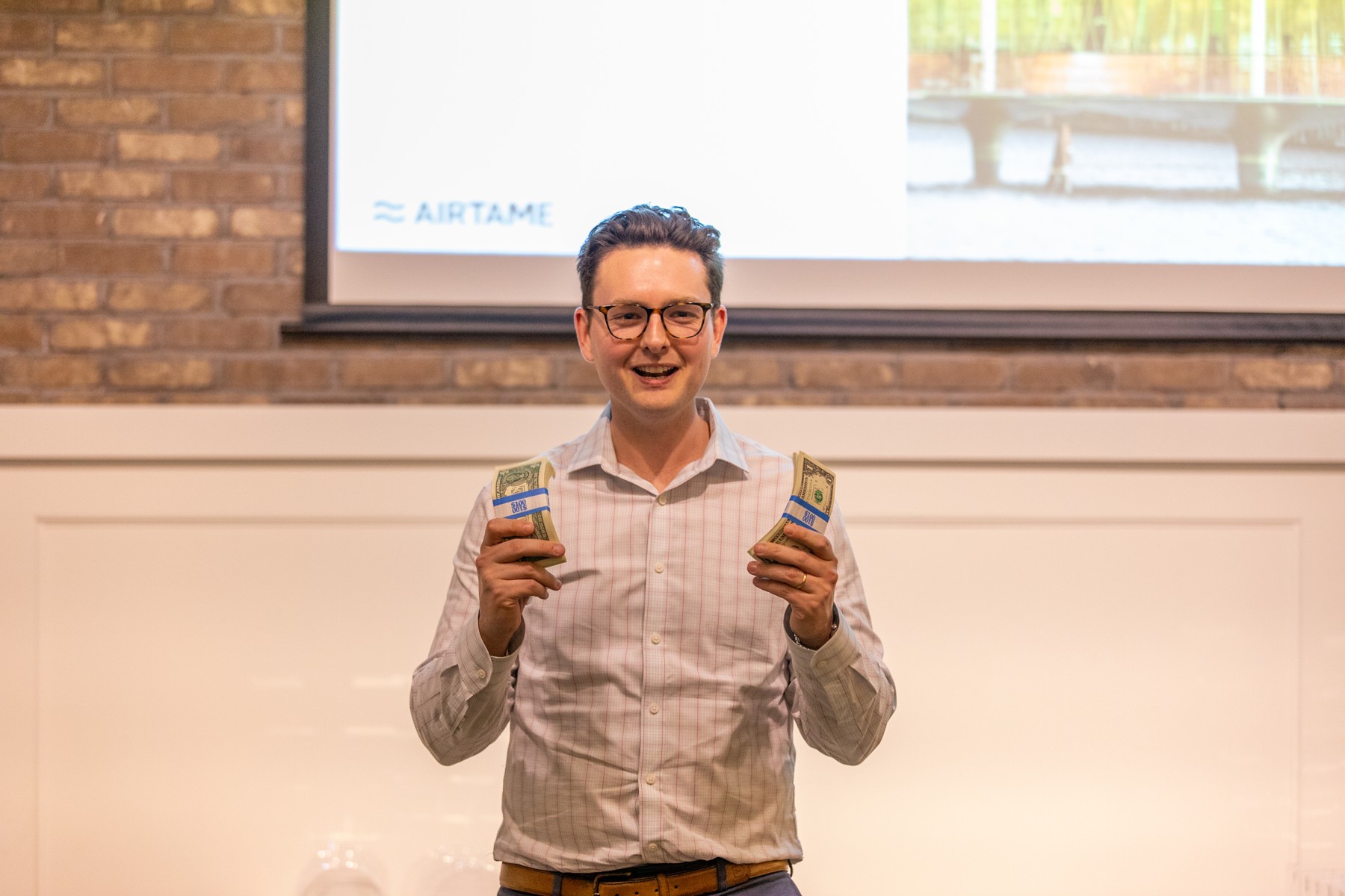 A man at a company event holds two bundles of banknotes in front of a presentation screen