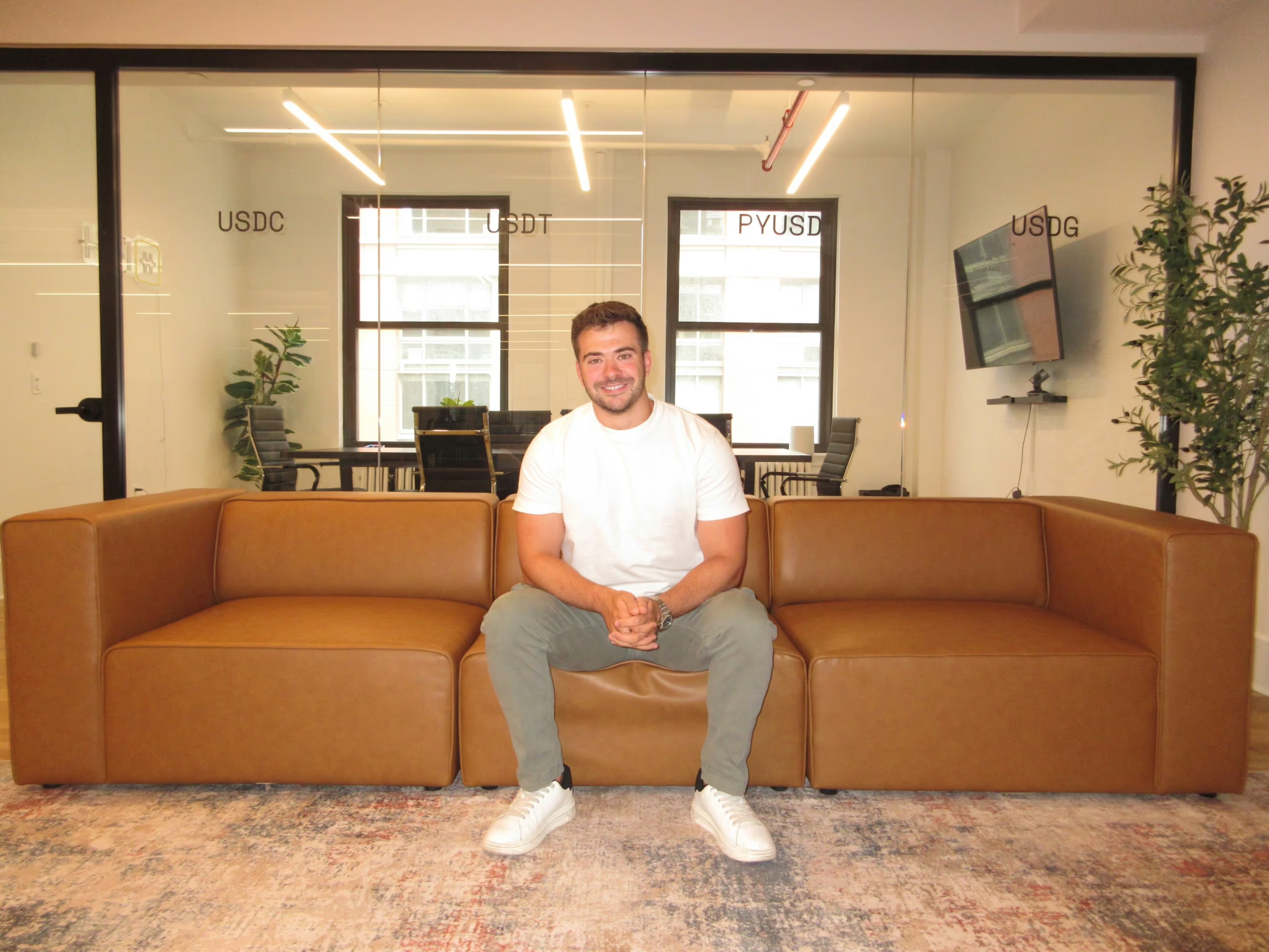 Zach Walsh sitting on a brown couch in the HIFI office, with stablecoin names on the meeting-room glass behind him