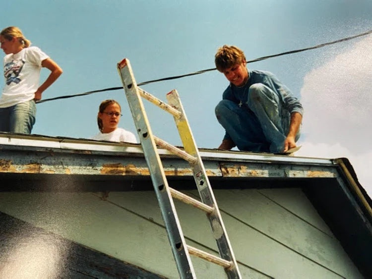 A younger Zack Abbott crouches on a roof beside a ladder during a building project