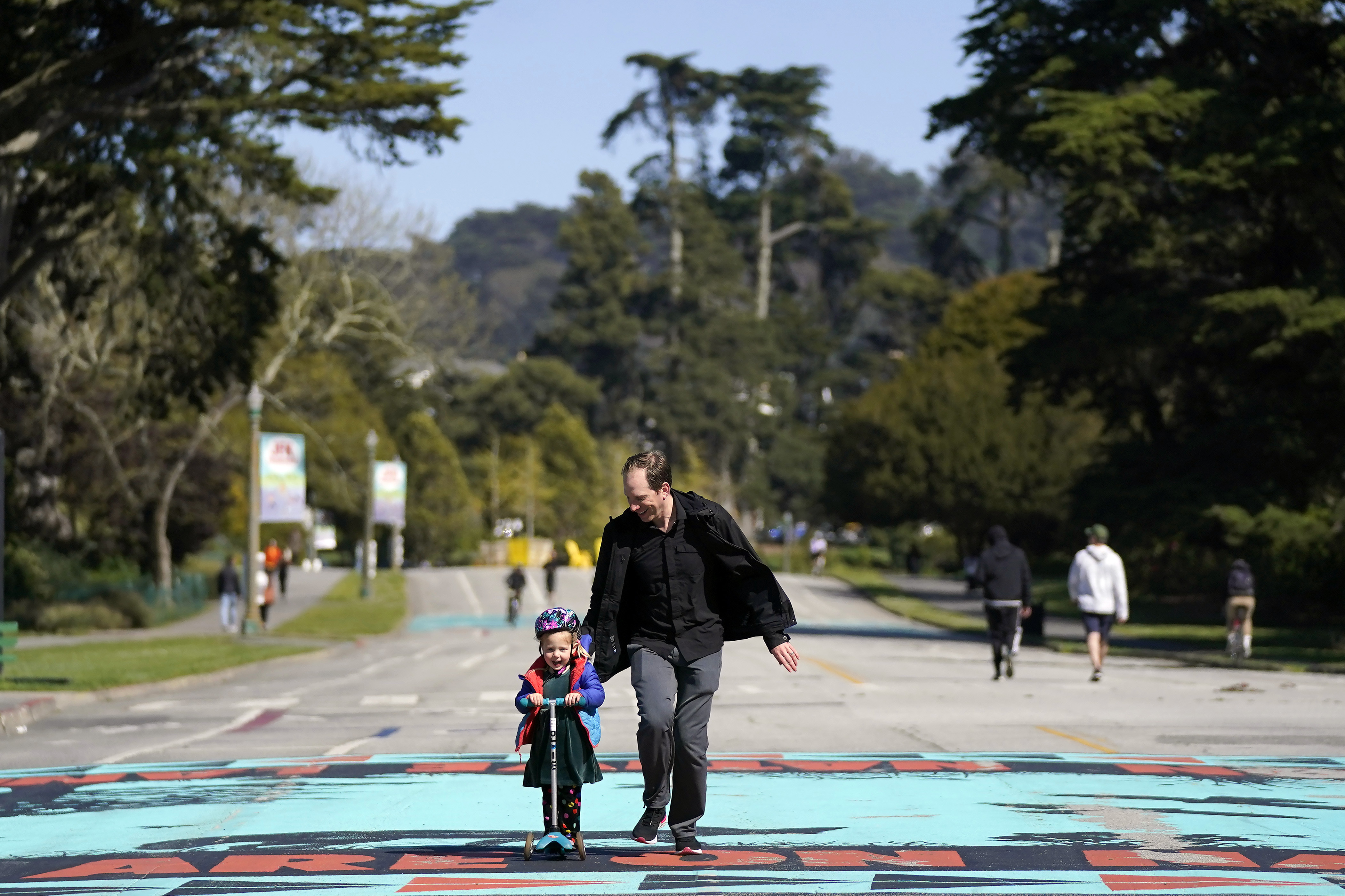 Zack Rosen running beside his daughter as she rides a scooter on John F. Kennedy Drive in Golden Gate Park