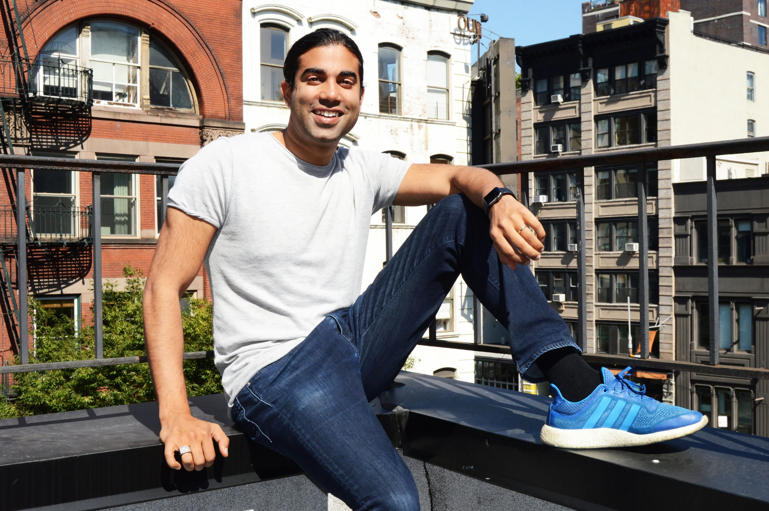 Zahir Dossa sitting on a New York rooftop in a gray T-shirt, jeans and blue sneakers