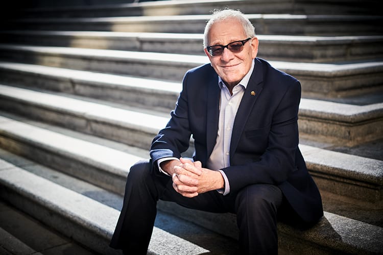 Zak El-Ramly seated outdoors on stone steps, wearing a dark suit and sunglasses