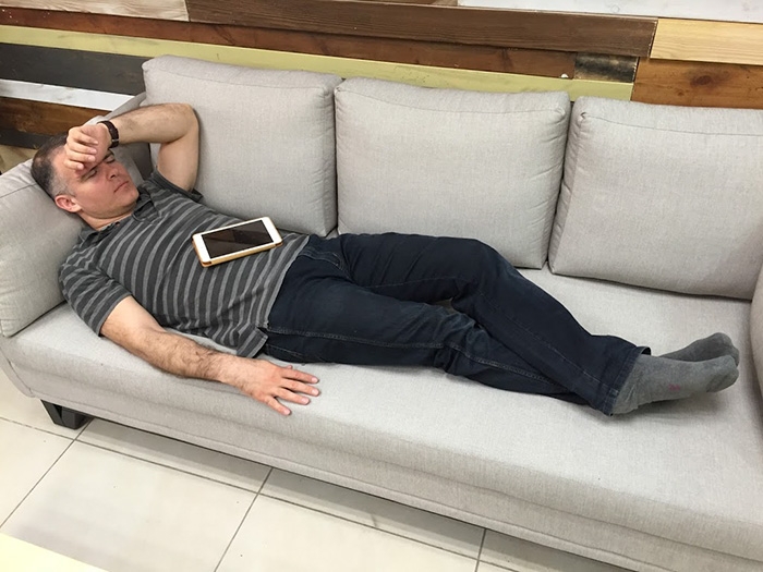 Zeev Farbman reclining asleep on a gray office sofa, with a tablet resting on his chest.