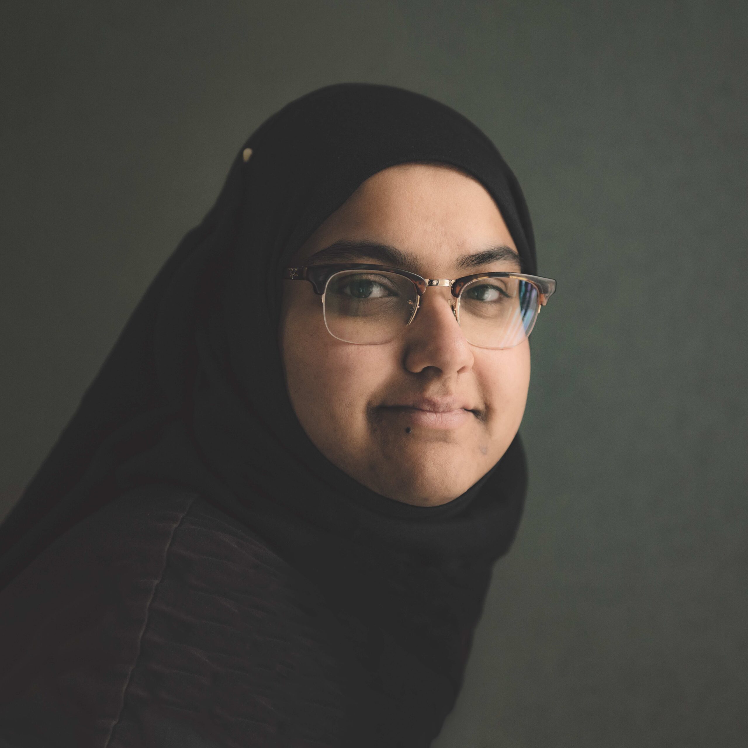 Zena Chaudhry wearing glasses and a black hijab in a press portrait against a muted green background