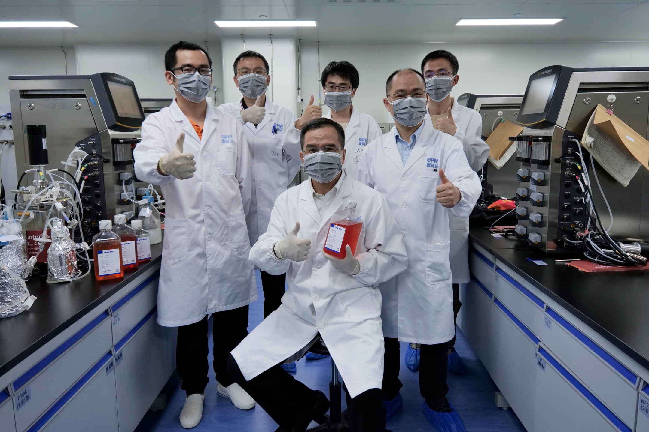 Zhihua Xiao seated with five OPM laboratory colleagues among bioprocessing equipment, all giving a thumbs-up