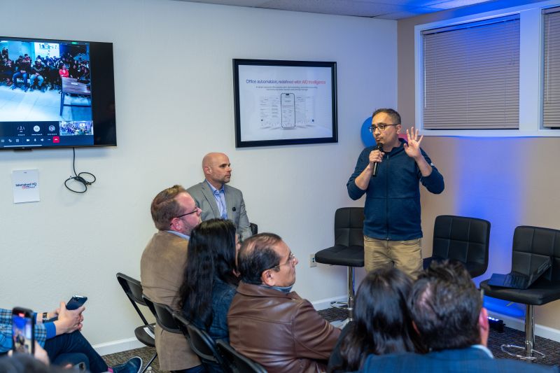 Zia Hasnain holds a microphone while speaking to a seated group, with a video meeting on a nearby screen.
