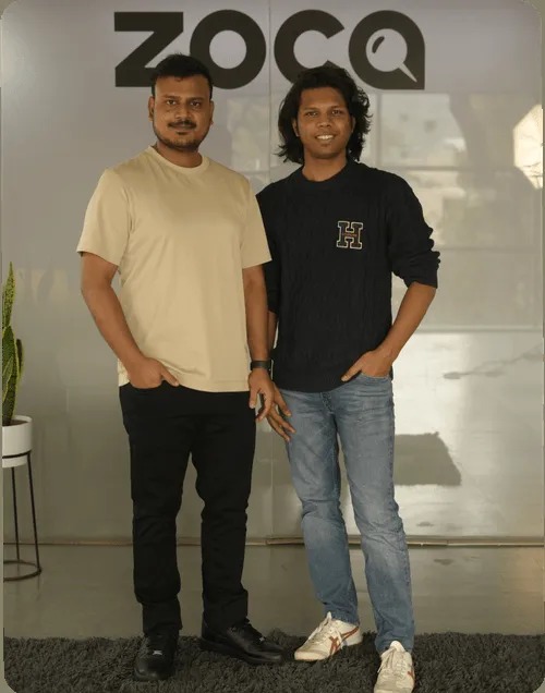 Two members of Zoca's founding team standing beneath the company logo
