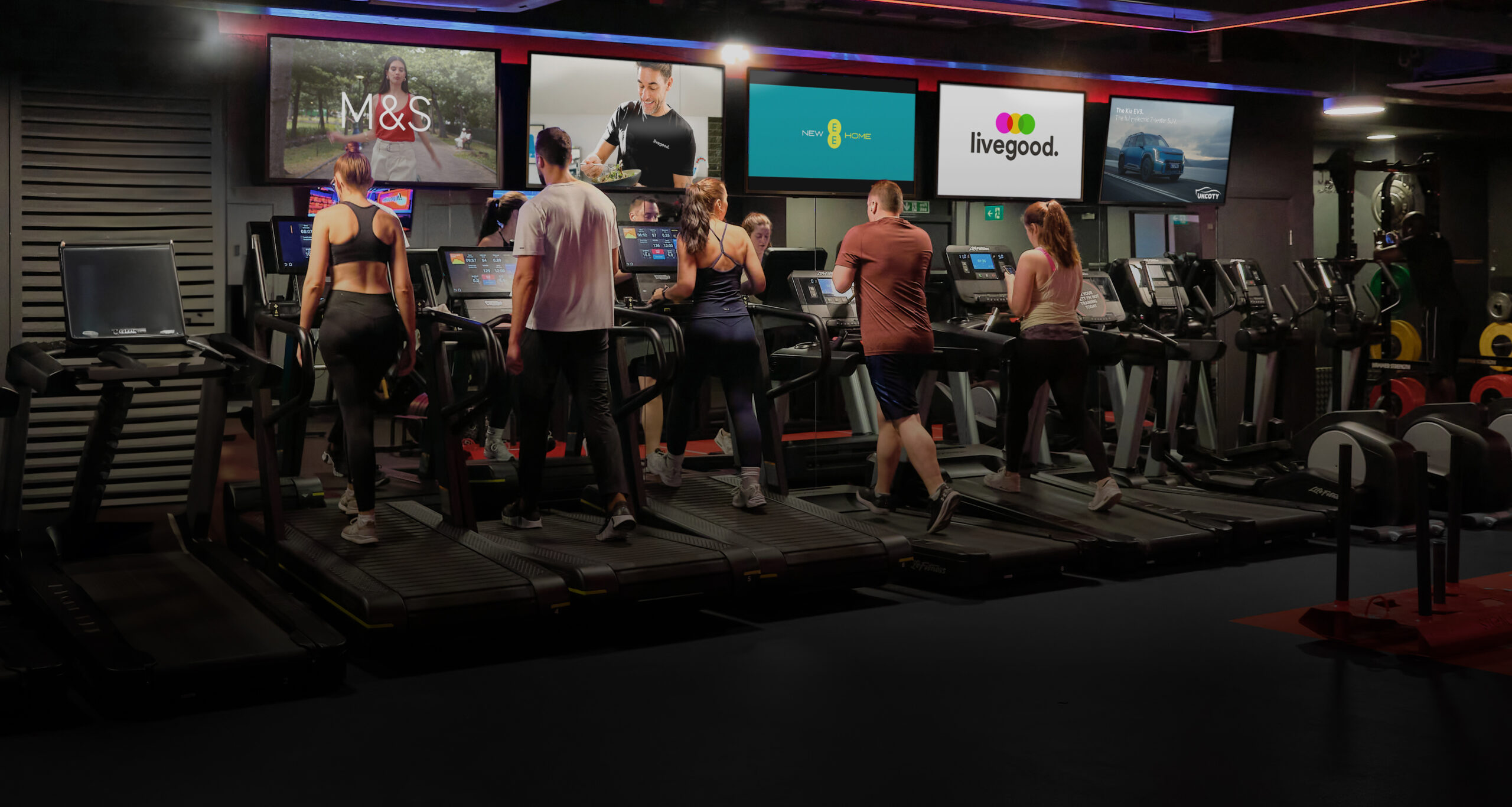 Gym members on cardio machines beneath screens displaying advertising and livegood wellness content