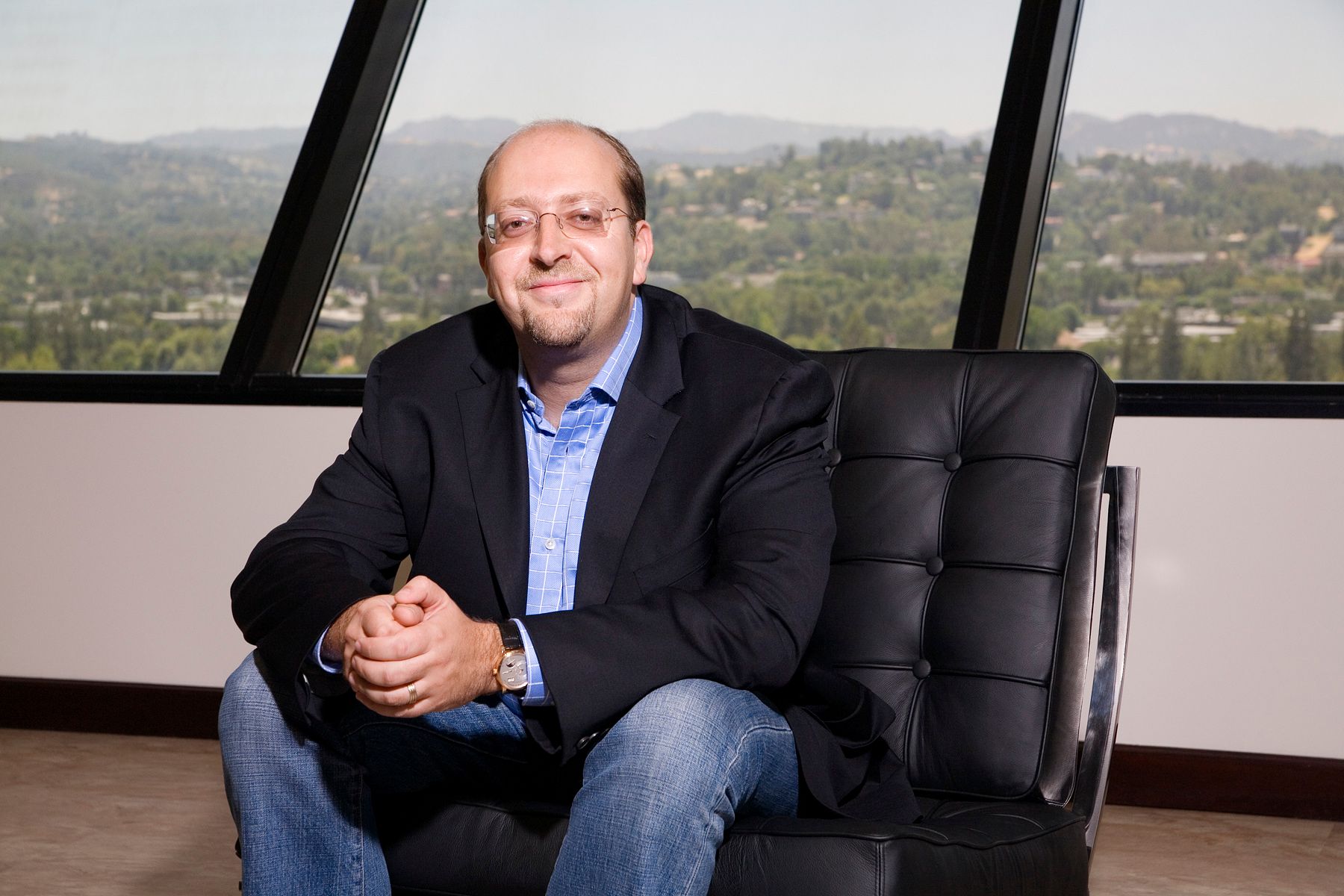 Zorik Gordon seated in a black chair beside office windows, wearing a blue shirt and dark blazer